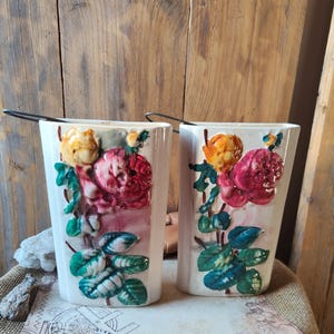 May include: Two white ceramic wall pockets with embossed floral designs. Each pocket features a bouquet of pink, yellow, and green flowers and leaves. The pockets have a rectangular shape and a metal handle. The background is a wooden wall.