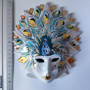 Peut inclure: Un masque en céramique blanc avec un motif de plumes de paon. Le masque présente une base blanche avec des trous noirs pour les yeux, des lèvres dorées et une coiffe de plumes détaillée dans des tons de bleu, vert et or. Le masque mesure environ 20 cm de hauteur.