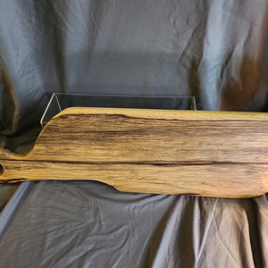 May include: A large, wooden cutting board with a unique, curved shape and a handle. The board has a natural wood finish with dark brown streaks throughout.