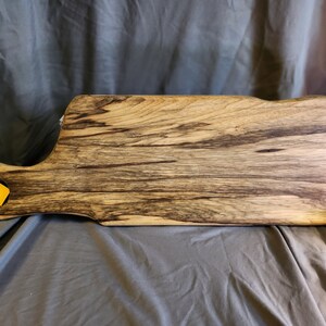 May include: A large wooden cutting board with a unique grain pattern and a handle. The board is dark brown with streaks of lighter wood. The handle has a small hole in it.