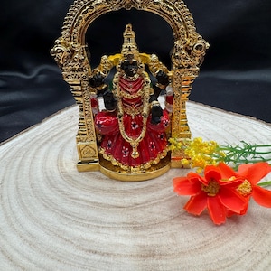 Op de afbeelding: Een gouden beeld van een hindoeïstische godheid, zittend op een troon met een rode en gouden mantel, in een gouden boog. Het beeld staat op een houten voet met een bloem.