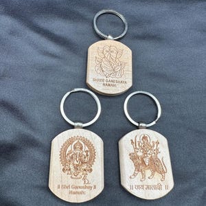 May include: Three wooden keychains with different Hindu deities engraved on them. The top keychain features Ganesha with the text "Shree Ganeshaya Namah". The middle keychain features Lakshmi with the text "II Shrid Ganoohsay II Namah:". The bottom keychain features Durga with the text "II Jay Matadi II".