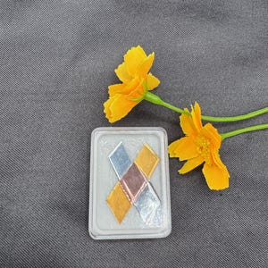 May include: A clear plastic case containing four small, square pieces of metal foil. The foil is gold, silver, copper, and a darker metal. The case is sitting on a gray fabric surface.