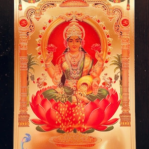 Op de afbeelding: Een gouden ingelijst beeld van Lakshmi, de hindoeïstische godin van rijkdom en voorspoed. Ze wordt afgebeeld zittend op een lotusbloem, met een pot met gouden munten in haar hand. Het beeld is omgeven door ingewikkelde gouden ontwerpen en de tekst "Jai Mata Di" is zichtbaar onderaan.