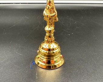 May include: A gold-colored bell with a detailed figure on top. The bell has a round base with a decorative border and colorful gemstones.