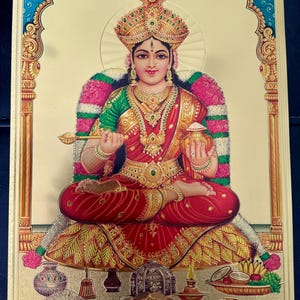 Puede incluir: Una imagen dorada de una diosa hindú sentada en un trono con un halo dorado. Lleva un sari rojo y verde, una corona y joyas. Sostiene una cuchara en una mano y una olla en la otra. Hay ofrendas de comida y fruta frente a ella.