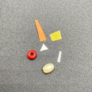 Puede incluir: Surtido de pequeños artículos decorativos coloridos, que incluyen una cuenta roja, una cuenta blanca, un rectángulo amarillo, un triángulo blanco, un trozo de papel rayado y un trozo de papel largo y naranja.