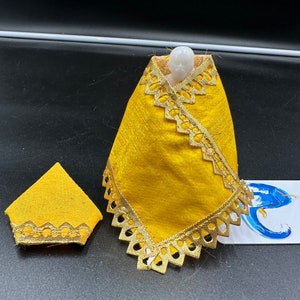 May include: A yellow fabric doll with gold trim. The doll is wearing a yellow fabric shawl with gold trim. The doll is sitting on a black surface.