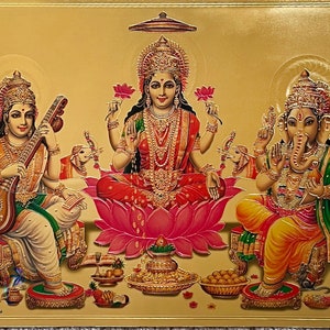 Op de afbeelding: Een gouden religieus beeld dat drie hindoe-goden voorstelt. De centrale figuur is een vrouw in een rode en gouden sari, zittend op een roze lotusbloem. De figuren aan weerszijden zijn een vrouw die een snaarinstrument bespeelt en een man met een olifantenkop. Het beeld is tegen een gouden achtergrond.