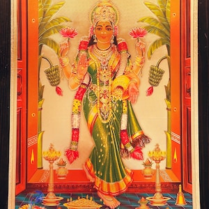 May include: A colorful painting of a Hindu goddess standing in a doorway. She is wearing a green sari and gold jewelry. She is holding a lotus flower in each hand. The painting is framed in gold.