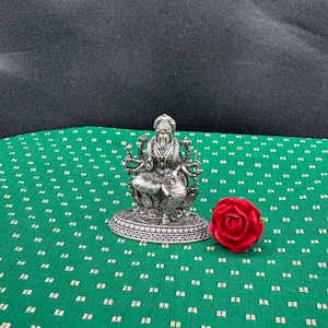May include: A detailed silver statue of a deity sits on a decorative base. A red velvet rose is next to the statue on a green patterned fabric. The background is a dark fabric.