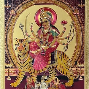 Op de afbeelding: Een kleurrijk schilderij van een hindoeïstische godin die op een tijger rijdt. De godin heeft meerdere armen en houdt verschillende wapens en symbolen vast. De tijger is afgebeeld in een gouden kleur en heeft een woeste uitdrukking. Het schilderij is geschilderd op een achtergrond van goud en bordeauxrood.