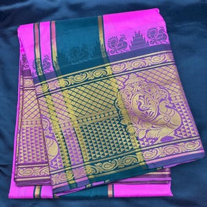 Op de afbeelding: Een roze en groene zijden sari met gouden borduurwerk. De sari heeft een geometrisch patroon en een bloemmotief. De sari is gevouwen en wordt getoond op een donkerblauw oppervlak.