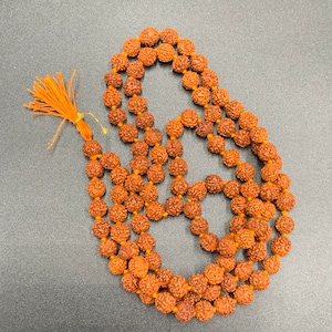 Puede incluir: Un collar de cuentas mala de color marrón con un borla de hilo naranja. El collar está hecho de 108 cuentas y se utiliza para la meditación y la oración.