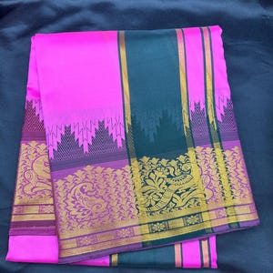 Op de afbeelding: Een opgevouwen roze sari met een donkergroene rand en gouden bloemen- en geometrische patronen. De sari heeft een brede donkergroene streep met gouden accenten en een roze en paarse rand. De stof is netjes op een donkere achtergrond gevouwen.