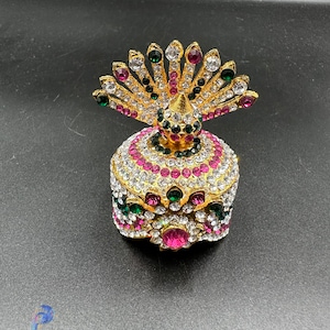 Op de afbeelding: Een goudkleurige, met juwelen bezette doos met een pauwenveermotief. De doos is ingelegd met heldere, roze en groene edelstenen. Het pauwenveermotief bevindt zich bovenop de doos.