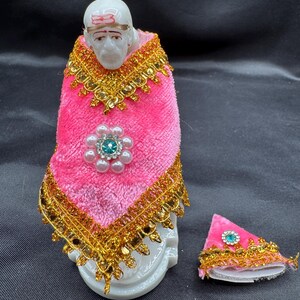 May include: A white statue of a religious figure adorned with a pink and gold embellished robe. The robe features a floral design with a blue gem. A matching hat is placed beside the statue.