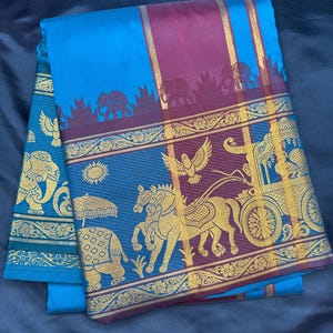 Op de afbeelding: Een blauwe en maroon zijden sari met een gouden rand. De sari heeft een herhalend patroon van olifanten, paarden en een wagen. De rand is versierd met bloemmotieven.