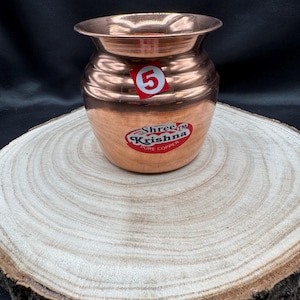 Op de afbeelding: Een koperkleurige pot met een brede opening en een afgeronde body, met horizontale ribbels. Een rood-witte sticker met het nummer "5" is op de pot bevestigd. De pot heeft een "Shree Krishna Pure Copper" sticker. De pot staat op een houten oppervlak.