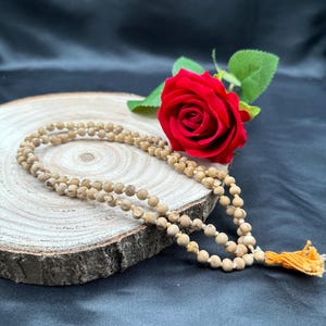 May include: A light brown beaded necklace with a yellow tassel rests on a wooden slice. A vibrant red rose with green leaves is placed next to the necklace against a dark background. The necklace is made of small, round beads.
