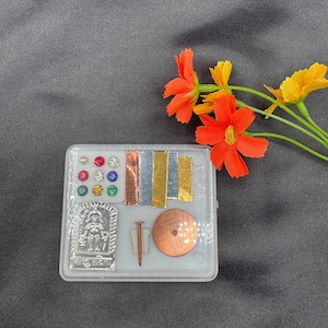 Op de afbeelding: Een klein, doorzichtig plastic doosje met verschillende charmes en snuisterijen. Het doosje bevat een zilveren charme met een klein figuurtje, een koperen schijf, een spijker en verschillende gekleurde edelstenen.