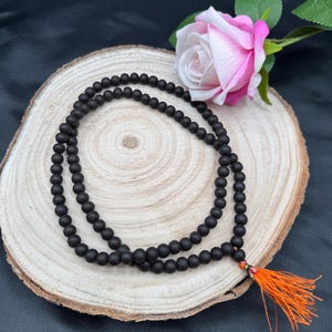 May include: A dark brown wooden mala bead necklace with an orange tassel. The necklace is on a wooden surface with a pink rose.