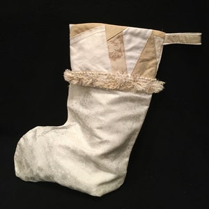 May include: A white Christmas stocking with a cream-colored patchwork top and a fluffy white trim. The stocking has a loop at the top for hanging.