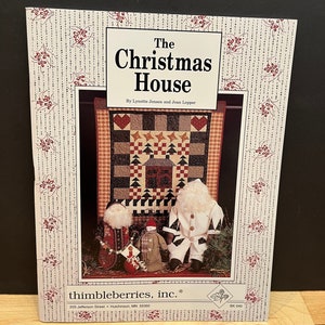 May include: A book titled "The Christmas House" by Lynette Jensen and Jean Lepper. The cover features a red and white patchwork quilt with a small house and Christmas trees. There are also three handmade dolls, one of which is a Santa Claus.