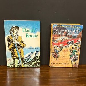 May include: Two vintage paperback books. The book on the left is titled "Daniel Boone" and features a man in a brown coat holding a rifle. The book on the right is titled "Tom Swift and His Triphibian Atomicar" and features a red car with a man and woman riding horses.