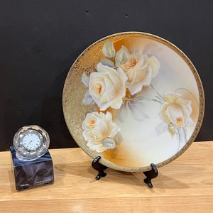 May include: A decorative plate featuring a floral design of white roses with yellow centers. The plate has a gold-colored rim and is displayed on a black stand. A crystal clock sits on a black box to the left of the plate.