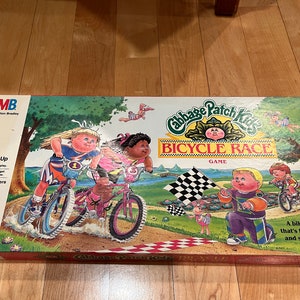 May include: A Cabbage Patch Kids Bicycle Race board game box with colorful illustrations of children riding bikes. The box features the game title "Cabbage Patch Kids Bicycle Race" and the text "A bike hike that's full of fun and surprises!"