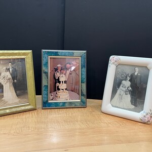 May include: Three framed photographs on a wooden surface. The frames are gold, teal, and white, each containing a black and white image of a wedding. The white frame has floral accents.