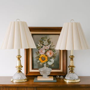 May include: Two matching table lamps with cream-colored pleated shades and brass bases. A floral painting and a decorative glass jar are in the background. The lamps are on a wooden surface.