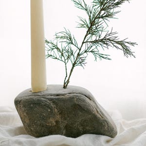 May include: A natural stone candle holder with a cream-colored candle and a small green plant. The candle holder is gray and sits on a white cloth. The candle is approximately 6 inches tall.