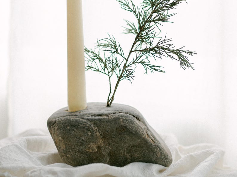 May include: A natural stone candle holder with a cream-colored candle and a small green plant. The candle holder is gray and sits on a white cloth. The candle is approximately 6 inches tall.