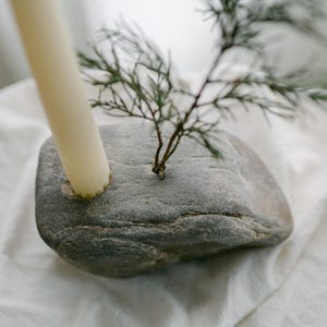 May include: A gray stone candle holder with a cream-colored candle and a small green plant. The candle is inserted into a hole in the stone. The plant is also inserted into the stone. The candle holder is on a white surface.