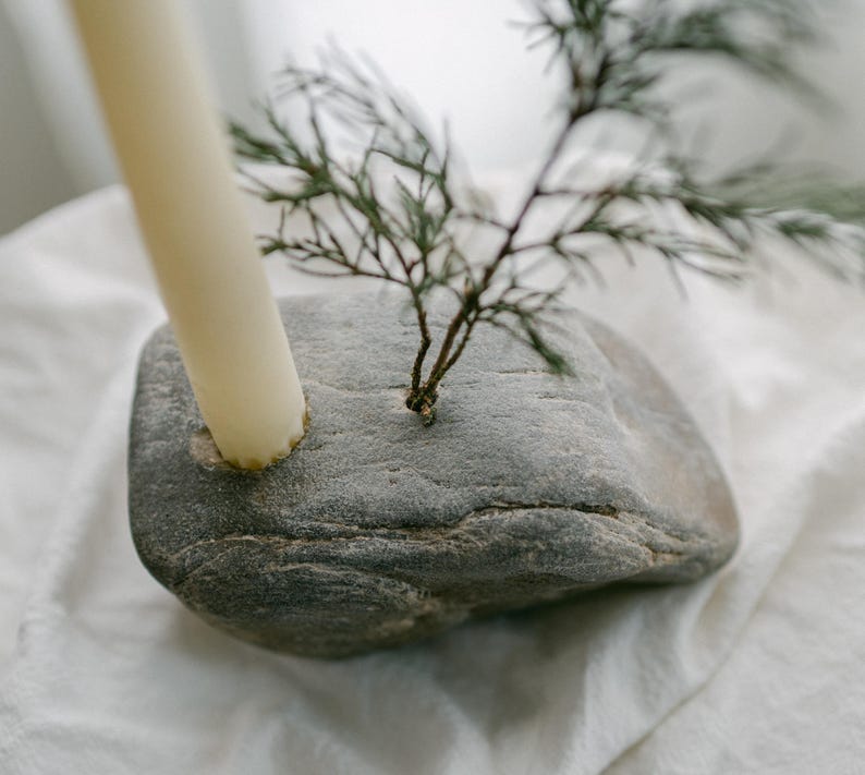 May include: A gray stone candle holder with a cream-colored candle and a small green plant. The candle is inserted into a hole in the stone. The plant is also inserted into the stone. The candle holder is on a white surface.