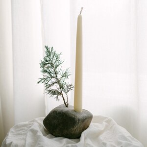May include: A cream-colored taper candle stands in a dark gray stone holder, with a small green sprig of foliage. The candle and holder are on a white, textured surface, with a white curtain in the background.