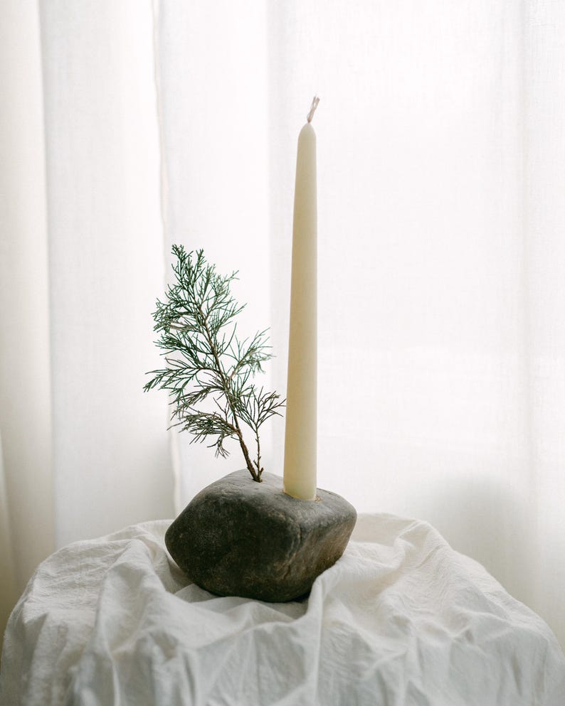 May include: A cream-colored taper candle stands in a dark gray stone holder, with a small green sprig of foliage. The candle and holder are on a white, textured surface, with a white curtain in the background.