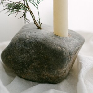 May include: A gray stone candle holder with a natural, textured surface. A beige candle and a small evergreen branch are inserted into the stone. The candle holder sits on a white, textured cloth.