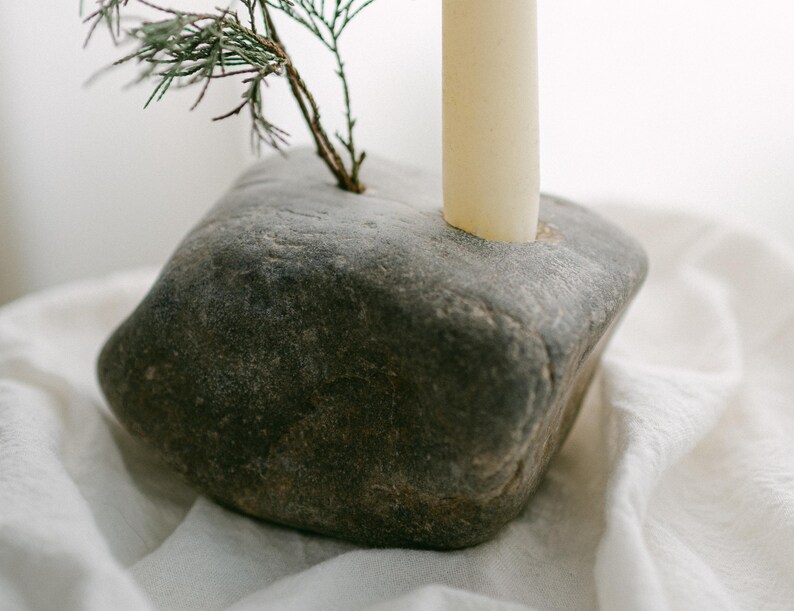 May include: A gray stone candle holder with a natural, textured surface. A beige candle and a small evergreen branch are inserted into the stone. The candle holder sits on a white, textured cloth.