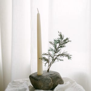 May include: A cream-colored taper candle stands tall in a gray stone holder, with a small green sprig of foliage adding a natural touch. The candle holder sits on a white cloth, creating a minimalist and elegant display.