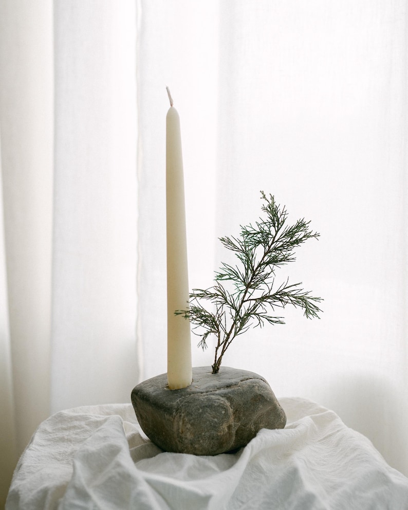 May include: A cream-colored taper candle stands tall in a gray stone holder, with a small green sprig of foliage adding a natural touch. The candle holder sits on a white cloth, creating a minimalist and elegant display.