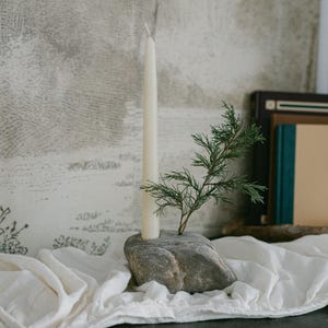 May include: A natural stone candle holder with a cream-colored taper candle and a small green sprig. The candle holder sits on a white cloth, with books in the background. The candle is approximately 10 inches tall.