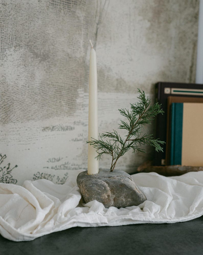 May include: A natural stone candle holder with a cream-colored taper candle and a small green sprig. The candle holder sits on a white cloth, with books in the background. The candle is approximately 10 inches tall.