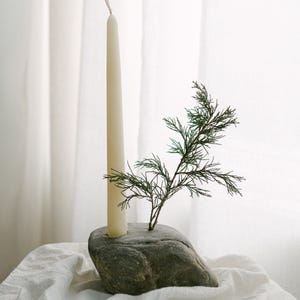 May include: A cream-colored taper candle in a gray stone holder, with a sprig of green foliage. The candle and stone are set on a white cloth, creating a minimalist, natural aesthetic. The candle is approximately 10 inches tall.