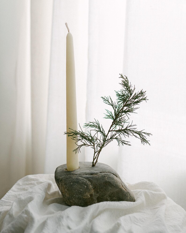 May include: A cream-colored taper candle in a gray stone holder, with a sprig of green foliage. The candle and stone are set on a white cloth, creating a minimalist, natural aesthetic. The candle is approximately 10 inches tall.