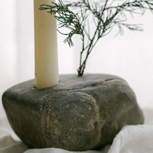 May include: A gray stone candle holder with a cream-colored candle and a small green plant. The candle holder is a natural, rounded stone with a hole for the candle. The plant adds a touch of greenery to the design.
