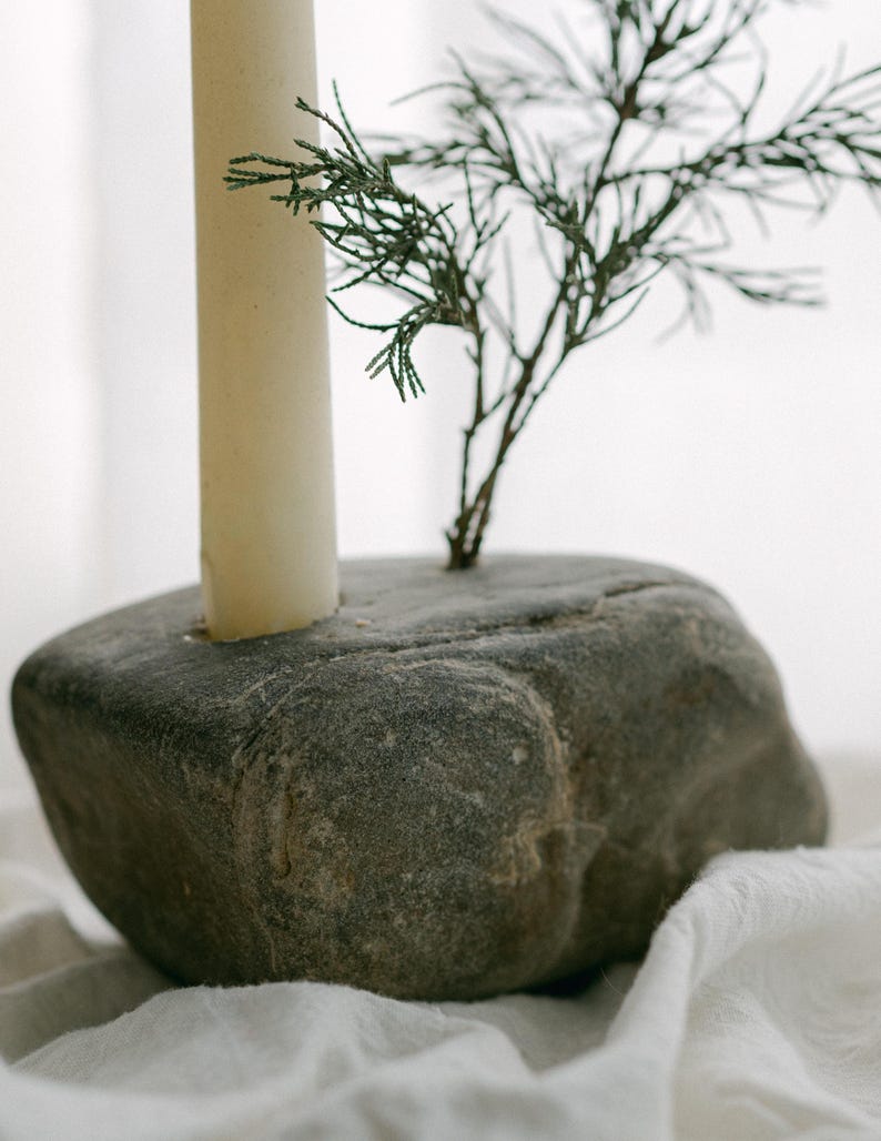 May include: A gray stone candle holder with a cream-colored candle and a small green plant. The candle holder is a natural, rounded stone with a hole for the candle. The plant adds a touch of greenery to the design.