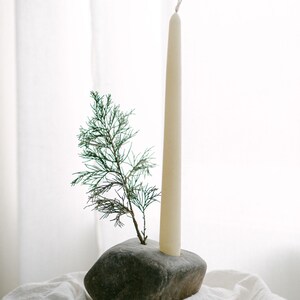 May include: A natural stone candle holder with a cream-colored taper candle and a small evergreen sprig. The candle holder is a dark gray color and sits on a white cloth. The candle is approximately 10 inches tall.